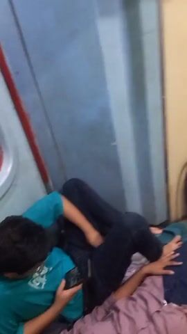 The video shows people sleeping on the floor of a train. In the first scene, a person wearing a blue shirt is sitting and looking at their phone. In the next scene, a fire extinguisher is visible. In the following scene, two people are sleeping on a blanket spread on the floor of a train compartment. A blue suitcase, a water bottle and a yellow bag are placed on the upper berth.