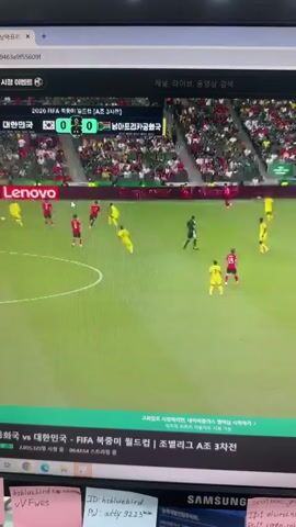 The video shows a computer monitor displaying a soccer match. The game is between South Korea and an opposing team, indicated by the scoreboard showing 0-0. The players are wearing red and yellow uniforms. Advertisements for Lenovo and "Smarter AI for all" are visible on the field. The bottom of the screen shows text in Korean, likely related to the match and broadcast information. Several sticky notes with handwritten notes and phone numbers are attached to the monitor and the area below it.
