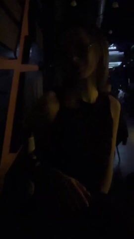 The video shows a young woman standing in a dark bar or club. She has long, light-colored hair and is wearing glasses and a black sleeveless top. The lighting is dim, but the woman is illuminated enough to see her face clearly. In the background, other people can be seen seated at tables, and there are various light fixtures. The woman looks directly at the camera with a slight smile. The camera angle appears to be from a handheld device. The video shakes slightly. There is music playing in the background. The overall atmosphere is casual and social.