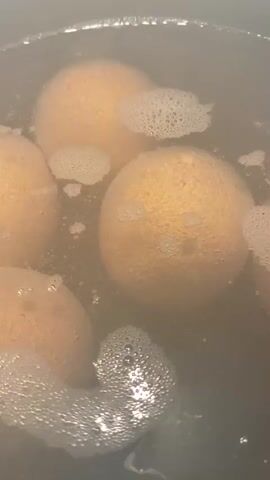 The video shows several whole oranges submerged in boiling water in a pot. The water is murky and bubbling, indicating it is hot. The oranges are round and have textured skin, appearing to be a typical orange color. Some of the oranges are partially obscured by foam and bubbles on the surface of the water. The lighting suggests an indoor setting, likely a kitchen. The camera is positioned above the pot, looking down at the oranges. The focus is on the oranges and the boiling water.