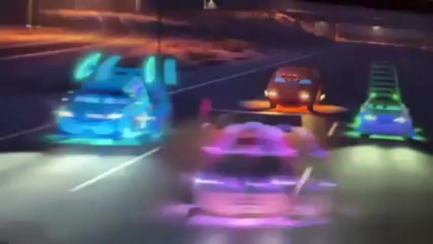 A TV screen displays a cartoon car race at night. There are four cartoon cars racing on a dark highway, with lights on.  The cars are brightly colored, in orange, blue, purple, and gray.  The camera angle is from behind the purple car, looking forward.  The background is blurred, with visible streetlights and some structures along the side of the road.  The TV is on a table covered with a floral patterned tablecloth, in front of a pink wall.