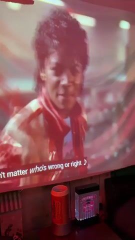 This media file captures a close-up projection of Michael Jackson singing the song "Beat It." The projection is displayed on what appears to be a large screen or banner, with a red and white color scheme dominating the image. Michael Jackson is seen in his iconic 1980s style, wearing a red and gold jacket with his signature curly hair. His facial expression is intense as he sings, with his mouth open and eyes focused.

Below the projection, several objects are visible. To the left, there is a cylindrical object, likely a can, with red and white branding that includes the word "Budweiser." Next to it, on the right, is a black rectangular light-up object with "Smirnoff" branding visible. The background behind these objects is blurred, but suggests an indoor setting with some striped patterns on the wall.

The overall mood is energetic and vibrant, characteristic of a music video performance. The lighting appears to be primarily from the projection itself, casting a red hue over the scene. The text displayed on the projection includes lyrics from the song: "doesn't matter who's wrong or right" and "Just beat it, beat it, beat it, beat it." The timestamp indicates the media is from early morning, around 00:00 to 00:04. While the specific location is Adachi, Japan, there are no overt visual cues in the image to confirm this detail beyond the provided metadata.