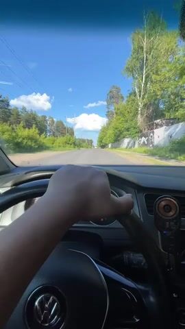 The video shows a first-person perspective of someone driving a car on a sunny day. The driver's hand is visible on the steering wheel, which has a Volkswagen logo. The road is paved and appears to be in a rural or suburban area, with trees and greenery on either side. There are power lines and poles visible along the left side of the road. A concrete wall with graffiti is on the right side. The sky is blue with some white clouds. The dashboard of the car is partially visible, including the speedometer and other instruments. There is a small, circular device mounted on the dashboard near the center. The car is moving forward.