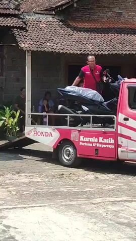 The video shows a man in a red vest and black pants standing next to a parked pink and white truck. The truck has the Honda logo on its side, and also writing that says "Kurnia Kasih Pertama & Terpercaya". The man is holding onto a black motorcycle that is on the back of the truck. The motorcycle is covered in plastic wrap. There are several people sitting on a porch in the background, watching the man with the motorcycle. There are also some potted plants on the porch. The house in the background has a tiled roof and brick walls. The ground in front of the truck is paved. The video appears to be taken in daylight.