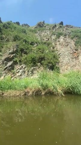 This is a first-person perspective video of someone kayaking on a river. The scene is set in a natural landscape with rocky, green hills on either side of the water. Tall green reeds line the banks of the river. The water is murky and reflects the sky and surrounding scenery.

The activity taking place is kayaking. The camera is positioned inside the kayak, showing the front of the bright red and yellow kayak. The kayaker's legs and feet are visible, resting in the kayak. A paddle is partially visible, extending out from the kayak. Two bright orange life jackets are also visible inside the kayak.

The time of day appears to be daytime, with a clear blue sky and bright sunlight. The weather seems to be warm and sunny, conducive to outdoor activities like kayaking. The location cues, such as the rocky hills and the vegetation, along with the provided context of Bikbovo, Russia, help establish the setting as a natural, possibly remote, river valley.

No text is explicitly visible in the video. The overall mood conveyed by the footage is one of peaceful exploration and enjoyment of nature.