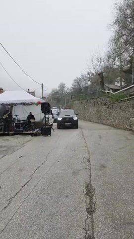 The video shows a street scene in a village on a cloudy day, likely overcast due to the even lighting. A black SUV slowly drives along the street, passing a live band that's set up under a white tent. The band consists of at least a saxophonist, drummer, and possibly others obscured by the tent and equipment. People are seated at picnic tables under a branded umbrella, and another person stands nearby. A dog trots in front of the SUV as it passes the band. The road is cracked and wet, suggesting recent rain or damp conditions. A stone wall borders one side of the road, and the background shows trees and houses, indicating a rural or village setting. Overhead power lines are visible. The overall atmosphere suggests a small community event or celebration in a rural area.