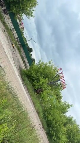 The video shows an outdoor scene with a dirt road in the foreground and lush green trees and vegetation. A large sign with red lettering is visible amongst the trees, although the text is partially obscured. A green fence runs along the left side of the frame, with some buildings or structures visible behind it. The sky is overcast and cloudy. A small yellow object is visible on the road in the distance. The camera is tilted, giving a diagonal perspective of the scene. The video is short, lasting only a few seconds.