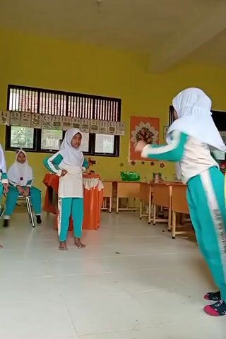 The video shows an indoor scene that appears to be a classroom. On the left, a young girl wearing a white hijab and a green and white tracksuit is seated on a chair. In the center, another young girl, also in a white hijab and a green and white tracksuit, stands with her hands on her hips, facing the viewer. To the right, a third young girl in a similar outfit is in motion, mid-jump with her legs bent and arms extended, suggesting she is performing a dance or a physical activity. She is facing away from the camera. Further to the right, a person partially visible, wearing a green and yellow shirt, is holding a white object, possibly a spray can, from which a stream of something is being expelled towards the performing girl. The room has yellow walls and a tiled floor. Desks and chairs are visible in the background, along with some decorative posters on the walls, including a tree motif. The lighting suggests daylight.