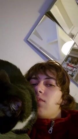 A young person with brown hair wearing a red zip-up jacket is holding a black and white cat on a gray surface. The person looks at the camera and puckers their lips as if to kiss the cat. The cat is mostly black with a white chest and paws, and it appears to be resting or sleeping. The background includes a wall with a framed picture and a light fixture. The camera angle is slightly tilted and close-up, focusing on the person and the cat.