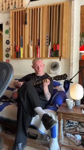 A young man with short, light blonde hair sits cross-legged in a comfortable chair, playing an acoustic guitar. He is dressed in a black t-shirt, black pants, white socks, and black shoes. His left arm has visible tattoos. The chair is covered with a blue and white patterned blanket. Behind him, there is a wooden structure on the wall with vertical slats, and a shelf above it holding various objects, including books and a small fan. To his right, a small wooden table holds a lamp, a mug, and various cables and electronics. An office chair is visible to the left, partially obscuring the view. The environment suggests a cozy, creative space, possibly a home studio.