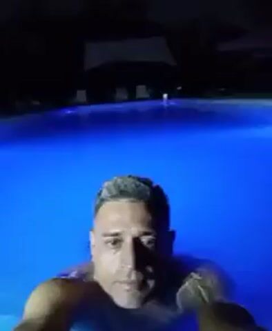 A medium shot shows a man treading water in a pool lit with blue light. The man has short, greying hair. He is shirtless. The pool is large. It is dark outside. In the background, several chaise loungers appear in the darkness near what may be a building. The man is looking directly at the camera and bobbing slightly in the water.