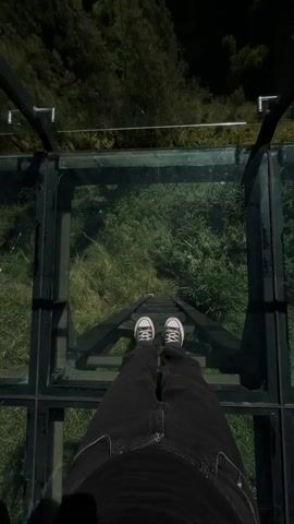 A first-person perspective video shows someone standing on a glass-floored observation deck. The deck overlooks a steep, lush green valley filled with trees. The person is wearing dark pants and black and white sneakers. The video pans slightly to show the expansive mountain landscape under a bright blue sky, with the sun shining.