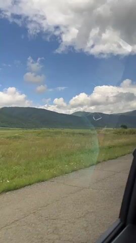 The video shows a view from inside a vehicle looking out at a rural landscape. A cracked asphalt road is visible in the foreground, with a grassy field extending beyond it. In the distance, a range of green, tree-covered mountains forms the horizon. The sky is a vibrant blue, dotted with fluffy white clouds. The sun appears to be shining, casting light on the scene. The overall impression is of a scenic drive through a natural environment. There are no people or specific actions depicted, other than the implied movement of the vehicle.