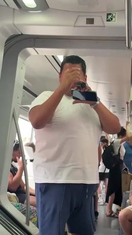 A man in a white t-shirt and blue shorts stands in a crowded train car, holding a smartphone and a small, orange object to his mouth. He appears to be singing or speaking into the phone, with his eyes closed and a passionate expression on his face. The train car is brightly lit, suggesting it is daytime. Other passengers can be seen in the background, some sitting and others standing. The overall atmosphere appears to be lively and energetic, possibly indicating a performance or a spontaneous moment of entertainment. The man's engagement with his audience, both on the phone and potentially within the train car, hints at a charismatic personality. The setting inside the train car, with its overhead compartments and seating, suggests a journey is underway. The visible signage with an orange pictogram and an arrow might indicate a safety instruction or an exit. Although the specific location is not explicitly visible, the mention of Torremolinos, Spain, places this scene within a popular tourist destination, possibly on a local or regional train. The man's performance, given the context of a public space, could be interpreted as a bold act of self-expression or a way to entertain fellow travelers.