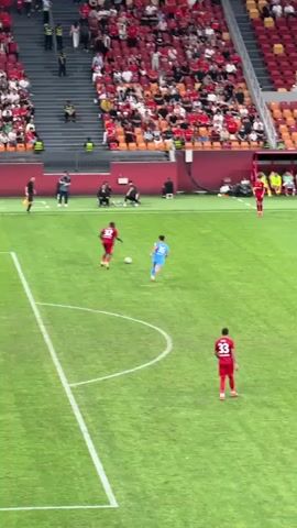 The video shows a soccer game in a stadium filled with spectators. Players wearing red uniforms are playing against players wearing light blue uniforms on a green grass field. The players are passing and running with the ball. The crowd is predominantly wearing red and waving flags, creating a lively atmosphere. There are also some yellow chairs on the sidelines near the field. The stadium has multiple tiers of seating and banners written in what appears to be Chinese characters. The camera moves rapidly, capturing both the action on the field and the energetic crowd.