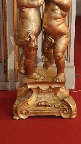 The camera slowly pans over a gilded architectural detail featuring two sculpted cherubic figures. The figures are depicted in a reclining position, one atop the other, appearing to support a section of the structure. The sculpture is finished in a golden tone and is positioned against a backdrop of pale gray and a segment of red wall. The detail appears to be part of a larger, perhaps antique, architectural element. The lighting is dim and emphasizes the texture of the gilded surface.