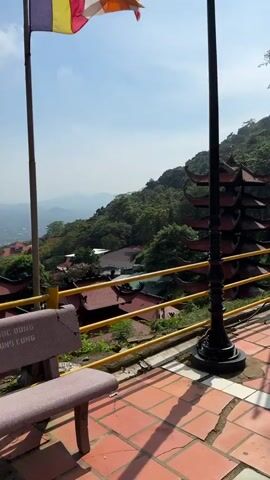 The video captures a scenic view from a high vantage point, overlooking a valley with scattered buildings and lush green hills under a bright, partly cloudy sky. In the foreground, a yellow railing runs horizontally, with a portion of a bench visible on the left, featuring faded lettering. A tall flagpole stands to the left, with the top section of a flag, displaying horizontal stripes of yellow, white, and red, visible. To the right, a decorative black lamppost is positioned near the railing. Below the railing, a complex of buildings with distinctive red, tiered roofs, characteristic of traditional Asian architecture, is spread across the hillside. Some of these structures appear to be pagodas. The dense foliage of trees and bushes covers the slopes of the hills, with a distant view of rolling mountains in the hazy background. The camera pans slightly, revealing more of the architectural layout and the expansive landscape.