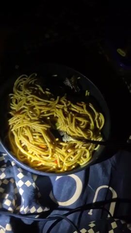 The video shows a plate of spaghetti with meat in a dark environment. There is also a bottle of Fanta and a blue container visible. The objects are on top of a checkered pattern fabric. The spaghetti is yellow and appears to have small pieces of vegetables mixed in. The lighting is poor, making it difficult to see details clearly.