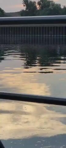 The video captures a tranquil scene of water, possibly a canal or river, in Klein-Krotzenburg, Germany. The water's surface is rippled, reflecting the muted colors of a sky that suggests either sunrise or sunset. Warm, golden hues mingle with softer grays and blues, indicating a gentle transition of light. A dark, horizontal railing, likely part of a bridge or walkway, dominates the foreground, partially obscuring the view of the water and the structure beyond.

In the background, a dark, imposing wall lined with vertical supports rises from the water, suggesting an engineered embankment or dam. The reflections on the water suggest there are trees or other structures on the opposite bank, though they are not clearly discernible. There are no people or distinct objects visible in the water or its immediate vicinity, contributing to the peaceful and somewhat solitary atmosphere. The overall impression is one of calm and quiet observation, with the interplay of light and water as the primary focus. The time of day is clearly either early morning or late evening due to the soft, reflective lighting. The weather appears calm, with the gentle ripples on the water indicating little to no wind. No text is visible in the media file.