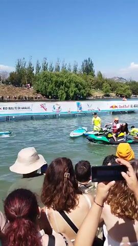 The video shows a crowd of people watching a paddleboard competition on a body of water.  The crowd is mostly facing the water, and many people are holding up their cell phones to record the event. In the water, a man in a yellow shirt is standing on a paddleboard and paddling towards a ramp. A jet ski is also in the water, carrying two people. A banner with “RED BULL TE DA AAAALAS” is visible behind the water. The environment includes trees, grass, and a clear blue sky.