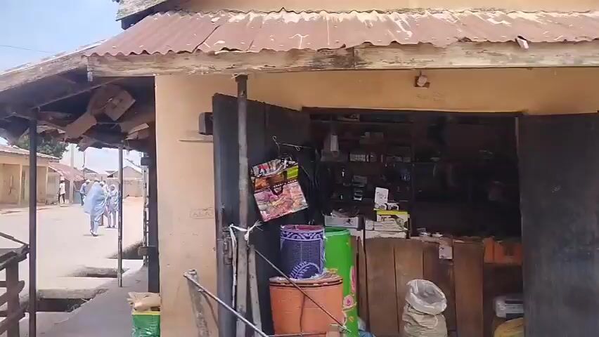 The video shows a dusty street lined with buildings, some made of mud brick with corrugated metal roofs, others appearing more permanent. A few people are visible walking along the street and near the buildings. One person is on a motorcycle. There are also some market stalls or shops with goods displayed. The sky is bright and cloudy. The overall impression is of a bustling, informal settlement or village.