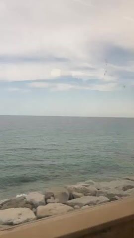 The video shows a view of the sea and the sky from inside a vehicle. The sea is calm with a slight turquoise hue. The sky is partly cloudy with streaks of white clouds against a light blue background. In the foreground, there is a rocky shore with large, light-colored stones. Part of a light brown surface, possibly a window frame or dashboard, is visible at the bottom of the frame. A thin tree with green foliage and a sign with yellow and blue elements appears briefly on the right side of the frame. The view suggests movement, possibly from a train or car traveling along the coast. There are no people visible in the video.