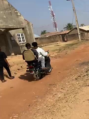 The video shows two people riding on a motorcycle down a red dirt road in a rural area. A person walks towards the camera on the left side of the road. Several houses, telephone poles, and a tall communication tower are visible in the background. The sky is blue.
