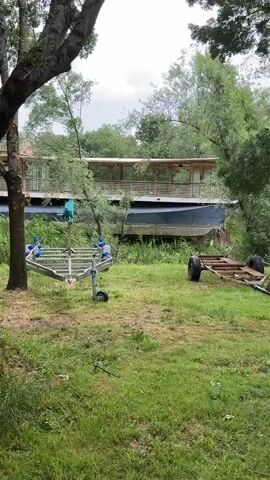 The camera is looking at an overgrown yard with green grass. There is a large boat with a wooden deck and a metal hull propped up on blocks and obscured by foliage. Two boat trailers, one silver and one rusted brown, sit in the yard. Trees surround the yard, creating a partial canopy above. The sky is overcast.