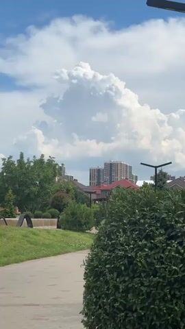 A video captured on a bright, cloudy day shows a view of a park and urban skyline. In the foreground, a pathway made of red and gray paving stones curves from the bottom left of the frame towards the center. To the left of the path is a strip of green grass, and a curved wooden structure, possibly part of a playground or a decorative element, is visible. Dominating the right side of the foreground is a dense, dark green bush. In the mid-ground, various trees and bushes are scattered, interspersed with the reddish-brown rooftops of houses. Beyond the houses, taller, modern apartment buildings with light-colored facades and visible windows rise up. The sky above is filled with dramatic, large white cumulus clouds, some of which are illuminated by sunlight, creating a contrast with the lighter gray tones of other clouds. The overall impression is a suburban or semi-urban landscape on a day with dynamic cloud formations. There are no people visible in the video.