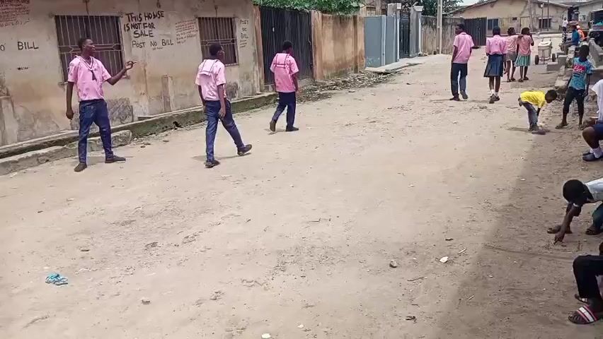 A group of young people, mostly wearing pink shirts and dark pants, are walking and standing in a dirt street in front of some buildings. Some of them appear to be school children. A few younger children are playing or sitting on the side of the street. The buildings are a mix of styles, with some appearing older and others more modern. There is some graffiti on one of the buildings. The environment appears to be a residential area.