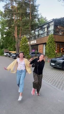 The video shows two women running down a paved path. The woman on the left is wearing a light yellow jacket over a white t-shirt with a floral pattern and blue pants. The woman on the right is wearing a black jacket and patterned pants. They are holding hands and appear to be laughing. The path is lined with trees and a building with a modern design is visible in the background. Several cars are parked on the side of the path. The lighting suggests it is daytime.
