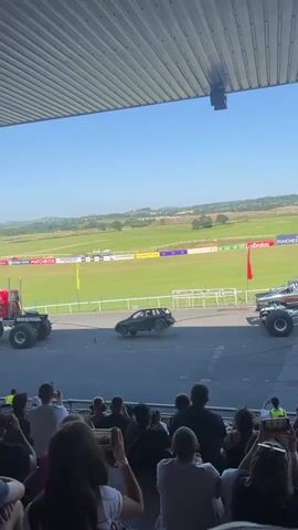 A crowd watches from bleachers as a monster truck drives over several cars. The monster truck is on the right side of the frame. Two crushed cars are in the center of the frame. A silver sedan is on the left side of the frame. A person in a red shirt is walking left to right in front of the silver sedan. A person in a yellow vest is standing on the right side of the frame. The background is a green field with a blue sky.