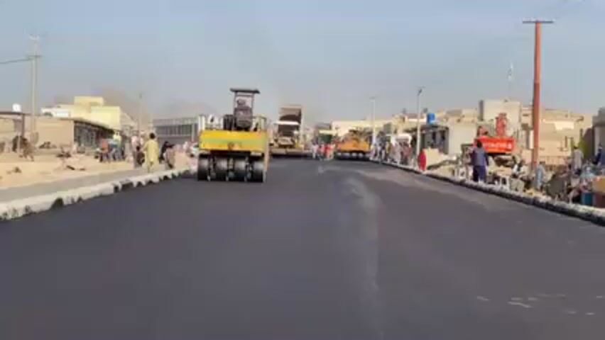 The video shows a recently paved asphalt road in a town, likely in a developing country. A yellow and black road roller is compacting the new asphalt. In the distance, a dump truck and another piece of construction equipment are visible. People are walking and standing on the sides of the road. Buildings, some appearing to be shops, line the street. Power lines and utility poles are present. In the background, there is a mountain. The sky is clear and the day appears to be sunny.