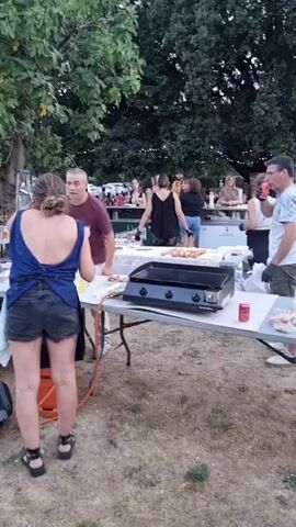 The video shows an outdoor gathering with several people and food service tables set up under trees. In the foreground, a woman with her back to the camera, wearing a blue top and black shorts, stands facing a table with a griddle. To her left, a man in a maroon shirt is interacting with someone off-camera. Behind them, other people are gathered around tables where food is being prepared and served. There are grills or griddles on the tables, along with various food items and drinks. A propane tank is visible next to one of the tables. The ground is covered with dry grass and dirt, and the overall atmosphere appears to be a casual outdoor event or barbecue.
