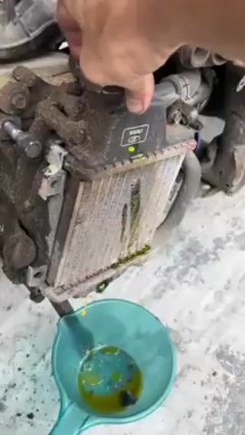 A close-up shot shows a dirty, green-stained radiator with a hand unscrewing the radiator cap. A green-yellow liquid is being drained into a turquoise container below. The radiator is connected to part of an engine. The setting seems to be outdoors on a gray concrete surface.