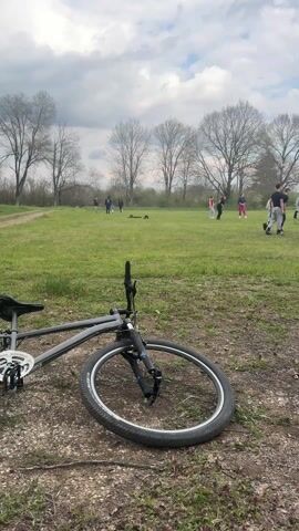 The video shows a grassy field, with a bike lying on the ground in the foreground. Further back, a group of people are playing volleyball. The camera pans to the left, revealing a dirt path, trees, and a gazebo. The sky is cloudy.