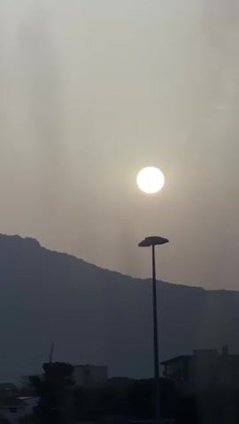 The video shows a hazy sky with the sun visible as a bright orb. A silhouette of a mountain range is in the background. In the foreground, a tall street lamp stands prominently. Below the lamp, indistinct buildings and trees are visible, suggesting an urban or suburban environment. The lighting is diffused, indicating either dawn, dusk, or a heavily overcast/hazy day. There are no people or distinct actions visible in the footage. The camera appears to be static or moving very slowly.