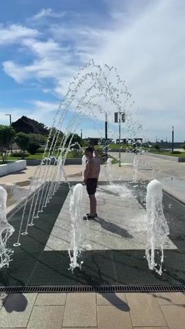 Two children are playing in an outdoor ground fountain display on a sunny day in Taman, Russia. The scene features multiple water jets shooting up from a paved surface, which is partially covered by metal grates.

A young girl, wearing a light-colored two-piece swimsuit with a patterned design, runs through the water. She appears joyful, smiling and at one point touching her mouth, perhaps due to water splashing on her face. Her hair is dark and pulled back into a ponytail.

In the background, a boy, wearing dark swim trunks, stands amongst the fountains, also getting wet. The sky above is blue with scattered white clouds, indicating clear weather. In the distance, there are some buildings or structures, green foliage, and lampposts, suggesting a public park or promenade area. The ground is made of light-colored paving stones. The overall atmosphere is playful and refreshing.