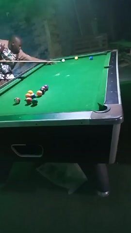 The video shows a man playing pool. He stands on the left side of the table, cue in hand, aiming at the racked balls. The pool table is a standard size with green felt and black rails. Various colored pool balls are arranged in a triangle at the center of the table, with the cue ball positioned near the opposite end.  The man wears a short-sleeved shirt with a light-colored floral pattern. The room is dimly lit, with a green hue illuminating the scene. Wooden pallets are stacked in the background, and a translucent plastic bag is visible underneath the pool table.