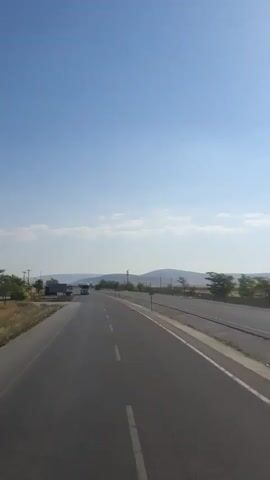 A highway stretches into the distance under a clear blue sky with scattered clouds. The road is paved and has a white dashed line marking the lanes. To the left of the road, there is a dry, grassy field with a few trees. To the right, there is a paved shoulder and then a fence. In the distance, there are hills and more trees. Two vehicles, possibly trucks or buses, are visible on the road ahead, moving away from the camera. The overall scene suggests a sunny day in a rural or semi-rural area.