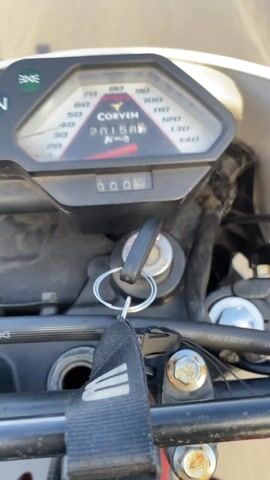 A first-person perspective video shows a person riding a Corven motorcycle on a highway during the day. The speedometer shows the motorcycle traveling at approximately 60-70 km/h. The odometer reads 20158 km. The key is in the ignition. The sky is blue with scattered clouds. Cars and trucks are visible on the highway ahead. A shadow of the person riding the motorcycle is visible.