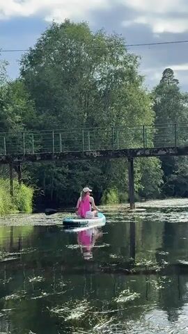 A person is sitting cross-legged on a paddleboard in a body of water. The person is wearing a pink top and a white hat. The paddleboard is in front of a bridge. The bridge is made of metal and has a green railing. The water is covered with green algae. Trees are visible in the background. The sky is cloudy.