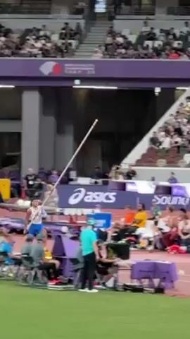 A medium, eye-level shot shows a pole vaulter clearing a bar in an outdoor stadium. An athlete with tanned skin, wearing a white tank top and blue shorts, runs toward the bar, places their pole into the launch box, and propels themselves over the bar. A large, seated crowd watches. The background is a crowded stadium and banners with sponsor logos such as ASICS. The bar is mounted on purple uprights. A cameraman is visible. The ground is a red track.