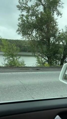The video shows the perspective of someone driving a car. The car is on a paved road, and there is a body of water to the left of the road, with trees and hills in the background. The sky is overcast. On the right side of the road, there are some signs and utility poles. Another car is visible driving in the opposite direction. The interior of the car, including the steering wheel and dashboard, is also visible. The time displayed on the dashboard is 17:49.