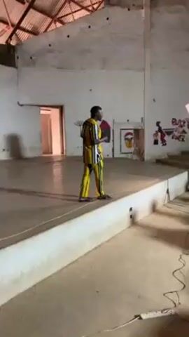 A man stands on a stage in a large room. The man is wearing a colorful striped shirt and matching pants. He has his hands clasped in front of him and appears to be speaking. The walls of the room are painted white, and there are some drawings on the wall. There is a doorway to the left of the stage. To the right of the stage is a set of stairs, and a light and cords are in the foreground. The room appears to be sparsely furnished.