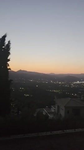 The video displays a nighttime aerial view of a coastal town with mountains in the background. The sky is a gradient of pale blue to pink, indicating twilight. Numerous lights dot the town, suggesting its population and activity. The foreground features a dark silhouette of trees and a building, partially obscuring the view. The camera seems to be panning slightly, offering a panoramic perspective of the landscape.