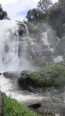 The video shows a majestic waterfall cascading down a rocky cliff face, surrounded by lush green foliage and trees. The water is white and frothy, creating a misty spray that hangs in the air. On the left side of the frame, a man stands near a wooden railing, holding up a phone to capture the scene. The camera angle is low, emphasizing the height and power of the waterfall. The sky above is overcast, with patches of blue peeking through the clouds. The overall atmosphere is serene and awe-inspiring, highlighting the natural beauty of the landscape.