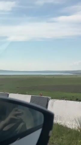 A car travels along a road. The camera is positioned inside the car, looking out the side window and at the side mirror. A concrete barrier runs along the edge of the road. Beyond the barrier is a vast green field with a body of water in the distance. The sky is blue with scattered clouds. A speed camera sign is visible in the field. A person's reflection can be seen in the side mirror.