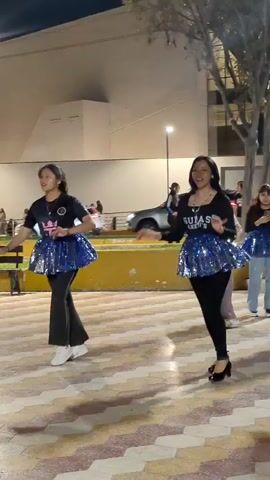 Two young women dance in unison at night in an outdoor, possibly public, square. The women are both wearing black shirts and pants, and sparkly blue miniskirts. The woman on the left has her hair in a ponytail and wears white sneakers, while the woman on the right has her hair down and wears black heels. They are both moving with energy, performing a synchronized dance. In the background, there is a building with lights, a small grassy area, and a street with cars and people. The pavement of the square is patterned in light and dark tones. The atmosphere appears lively and festive.