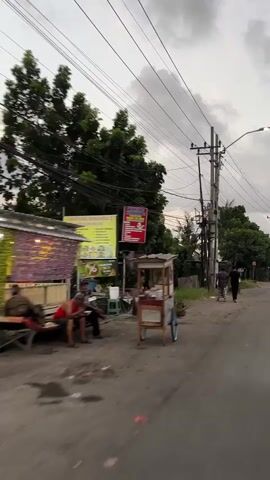 The video begins with a dynamic perspective of a street scene, possibly captured from a moving vehicle. The camera pans slightly to the right, revealing a roadside with several elements. On the left, a small stall or shop is visible, with purple and white striped fabric draped over its structure. Several people are seated on a wooden bench outside this establishment, appearing to be resting or socializing. One individual is wearing a red tank top. Adjacent to the stall, a yellow sign with text and images is mounted on a post. Further down the street, a small mobile food cart with wooden and metal components is parked on the roadside. The street itself is paved with asphalt, and there are patches of dirt and debris. Overhead, a complex network of electrical wires crisscrosses the sky, supported by wooden and concrete poles. Lush green trees line the right side of the road, creating a natural boundary. In the distance, a few figures are walking along the street, adding a sense of ongoing activity. The lighting suggests it is either early morning or late afternoon, with diffused light and a somewhat overcast sky. The overall impression is of a casual, everyday street environment in a tropical or subtropical location.